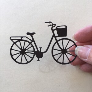 Puede incluir: Silueta negra de una bicicleta con una cesta en el manillar. La bicicleta tiene un dise&ntilde;o cl&aacute;sico con un cuadro curvo y ruedas de radios.