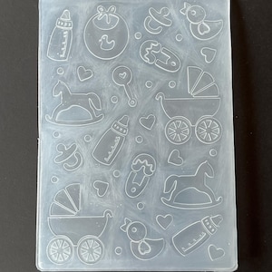 May include: A clear, rectangular silicone mold featuring baby-themed designs. Outlines of a baby bottle, bib, pacifier, duck, baby carriage, rocking horse, and hearts are visible. The mold is designed for crafting or baking.