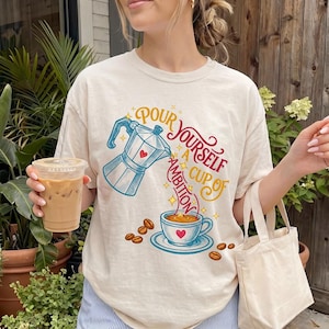 May include: A cream-colored t-shirt featuring a graphic of a moka pot pouring coffee into a cup. The text reads, "Pour yourself a cup of ambition," with small red hearts and yellow stars surrounding the coffee-themed illustration.
