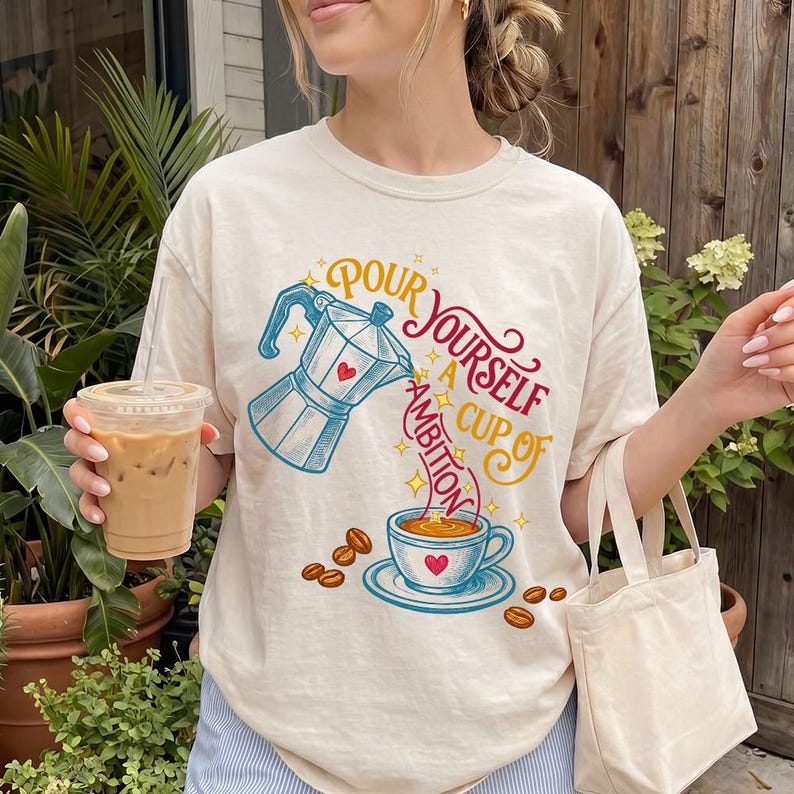 May include: A cream-colored t-shirt featuring a graphic of a moka pot pouring coffee into a cup. The text reads, "Pour yourself a cup of ambition," with small red hearts and yellow stars surrounding the coffee-themed illustration.