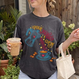 May include: A charcoal gray crew neck t-shirt featuring a graphic print of a blue moka pot pouring coffee into a blue cup. The text reads "POUR YOURSELF A CUP OF AMBITION" in yellow and red script. Scattered coffee beans and small stars surround the design.