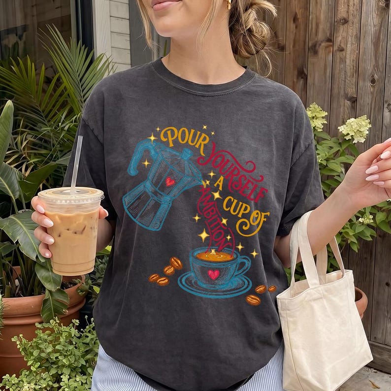 May include: A charcoal gray crew neck t-shirt featuring a graphic print of a blue moka pot pouring coffee into a blue cup. The text reads "POUR YOURSELF A CUP OF AMBITION" in yellow and red script. Scattered coffee beans and small stars surround the design.