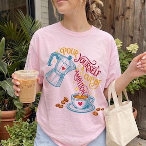 May include: A light pink t-shirt featuring a graphic of a blue moka pot pouring coffee into a cup. The text reads "POUR YOURSELF A CUP OF AMBITION" in red and yellow script. Scattered coffee beans and small yellow stars surround the central design.