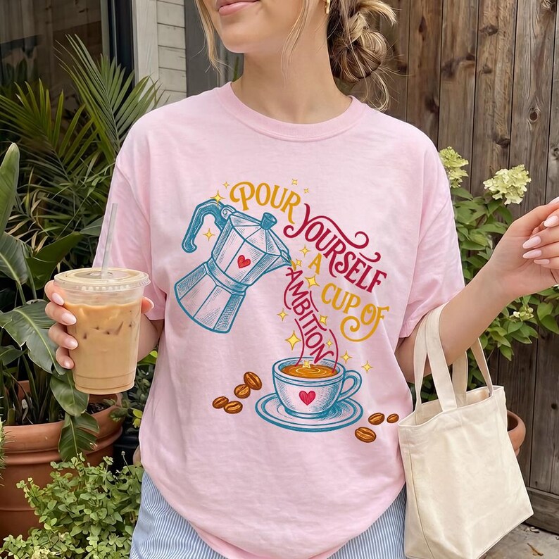 May include: A light pink t-shirt featuring a graphic of a blue moka pot pouring coffee into a cup. The text reads "POUR YOURSELF A CUP OF AMBITION" in red and yellow script. Scattered coffee beans and small yellow stars surround the central design.