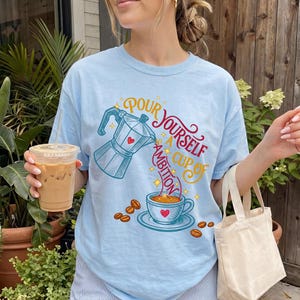 May include: A light blue t-shirt featuring a graphic of a moka pot pouring coffee into a cup with the text "Pour yourself a cup of ambition" in red and yellow script. The design includes small yellow stars and scattered brown coffee beans.
