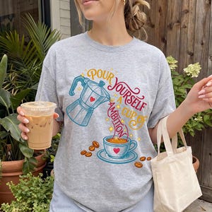 May include: A heather gray t-shirt featuring a graphic of a blue moka pot pouring coffee into a blue cup. The text reads "POUR YOURSELF A CUP OF AMBITION" in red and yellow script. Scattered brown coffee beans and small yellow stars surround the design.