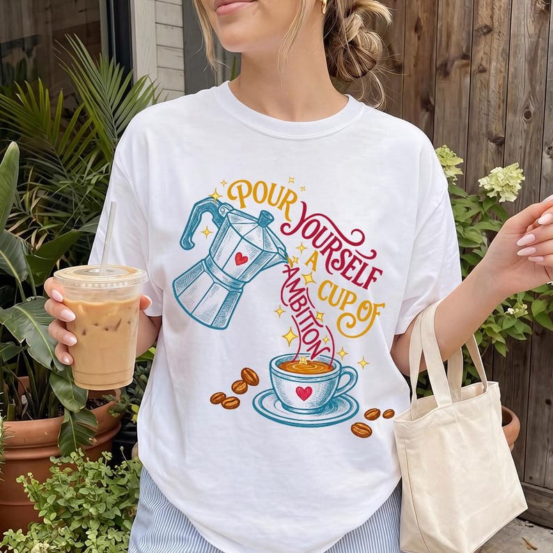 May include: A white t-shirt featuring a graphic of a blue moka pot pouring coffee into a cup with the text Pour Yourself A Cup Of Ambition in red and yellow, surrounded by small coffee beans and yellow stars, with a clear plastic cup of iced coffee.