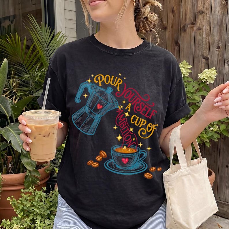 May include: A black t-shirt featuring a graphic print of a blue moka pot pouring coffee into a blue cup. The text reads, "Pour yourself a cup of ambition," with small yellow stars and scattered coffee beans surrounding the central design.