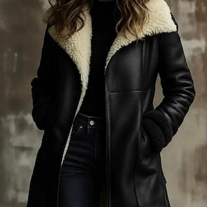 May include: A black coat with a smooth exterior finish and a wide, cream-coloured shearling lapel. The coat features long sleeves with matching black shearling cuffs, a straight hemline, and two deep front pockets, worn over a black shirt and jeans.