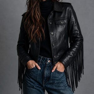Puede incluir: Una chaqueta de cuero negro con largos flecos en las mangas y la espalda, cuello clásico, cierre frontal de botones y dos bolsillos en el pecho con solapas abotonadas. La chaqueta se lleva sobre un suéter de cuello alto negro y vaqueros azul oscuro.