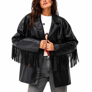 Puede incluir: Un blazer de piel sintética negra con mangas largas con detalles de flecos, cuello de solapa con muescas y dos botones frontales. La chaqueta se lleva sobre una camiseta blanca con un estampado gráfico y pantalones vaqueros grises.