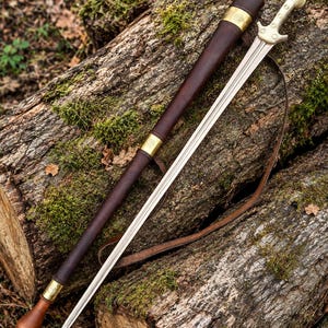 Puede incluir: Una espada de doble filo con hoja metálica, pomo de color claro y guarda. La espada va acompañada de una vaina de cuero marrón oscuro con detalles en latón y una larga correa de cuero para transportar el arma de forma segura.