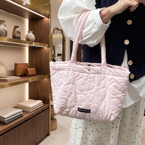 May include: A light pink quilted tote bag featuring an all-over heart pattern, two padded shoulder straps, a gold-toned snap button closure at the top, and a small rectangular black fabric label with the word ROMANCE printed in white text.