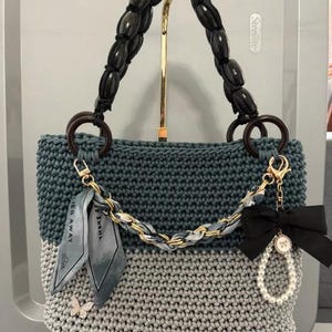 May include: A two-toned crochet handbag featuring a dark teal upper section and a light gray lower section. The bag has a dark wooden bead handle, a gold chain strap with a gray ribbon, a black bow, a pearl loop, and a small butterfly charm.