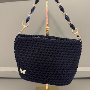 May include: A dark navy blue crocheted handbag featuring a textured stitch pattern and a small silver-toned butterfly charm on the lower left corner. The bag includes a decorative handle made of dark blue oval beads linked by gold-toned metal rings.