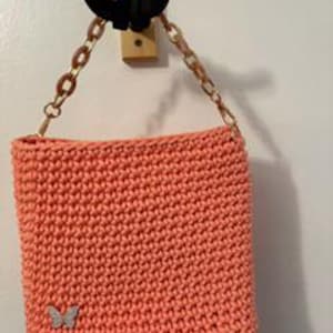 May include: A peach-colored crochet handbag featuring a rectangular body with a dense, textured stitch pattern. The bag includes a detachable gold-toned chain strap with translucent peach-colored links and a small silver butterfly charm.
