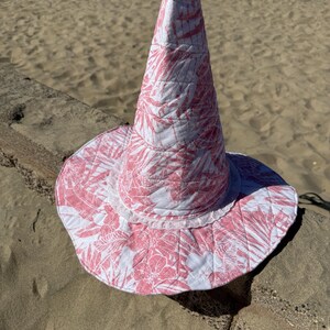 May include: A quilted cone-shaped hat featuring a pink and white tropical floral pattern. The hat has a wide, circular brim and a white lace trim detail encircling the base of the tall, pointed crown, constructed with visible stitching throughout.