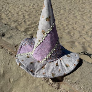 May include: A conical hat featuring a patchwork design of light gray fabric with small orange floral patterns and textured purple fabric. The sections are joined by light green rickrack trim, and the entire piece has visible quilted stitching.