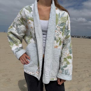 May include: A quilted jacket featuring a light green and white floral pattern on the sleeves and back, contrasted with a light blue and white pinstriped fabric on the collar, cuffs, and front pockets. The garment has a shawl collar and long sleeves.