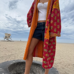 May include: A long, open-front quilted duster coat featuring a vibrant red and orange color palette with a repeating yellow paisley pattern. The garment has long sleeves, a hood, and visible white stitching throughout the quilted fabric.
