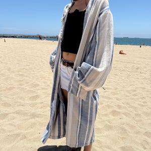 May include: A long-sleeved, open-front robe featuring a woven texture with vertical stripes in shades of blue, white, and beige. The garment has a hood, wide sleeves, and a relaxed, knee-length silhouette, worn over a black top and white shorts.