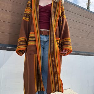 May include: A long, open-front brown coat featuring wide, vertical stripes in shades of orange, yellow, and dark brown along the lapels and sleeves. The garment has a loose, relaxed fit with long sleeves and a textured, woven appearance throughout.
