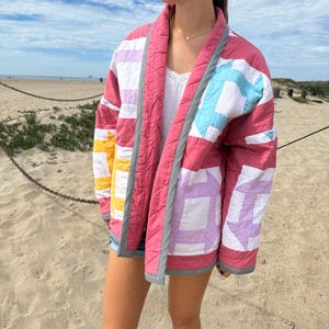 May include: A quilted jacket featuring a patchwork design with blocks of pink, white, yellow, and light blue fabric. The garment has a shawl collar and edges trimmed with a black and white checkered pattern, showcasing a stitched diamond texture.