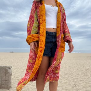 May include: A long, open-front quilted robe featuring a vibrant patchwork design in shades of red, orange, and green with intricate paisley and floral patterns. The garment has long sleeves, a wide mustard-yellow trim, and two large front pockets.