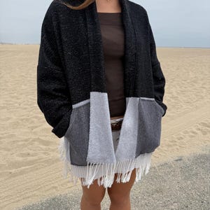 May include: An open-front cardigan featuring a black upper section and a color-blocked lower section in light gray and dark gray. The garment has long sleeves, two front pockets, and a white fringed hemline along the bottom edge.