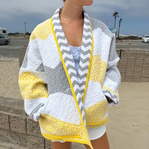 May include: A quilted jacket featuring a geometric pattern in yellow, white, and gray. The garment has a V-neckline, long sleeves, and a front border with a gray and white chevron print. The fabric displays a textured, stitched finish throughout.
