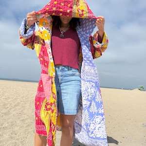 May include: A long, hooded coat made from quilted fabric featuring a patchwork of vibrant colors including magenta, yellow, and white with floral and geometric patterns. The interior lining displays a contrasting light blue and white botanical print.