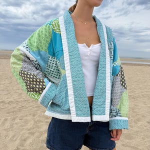 May include: A quilted jacket featuring a patchwork design of various blue and green patterned fabrics. The garment has long sleeves, a white trim along the front opening and cuffs, and a relaxed fit. It is worn over a white tank top and denim shorts.