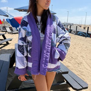 May include: A quilted jacket featuring a patchwork design with white, purple, and floral patterned fabric blocks. The garment has a solid purple wide collar, long sleeves with cuffs, and two front pockets, worn over a white graphic t-shirt.