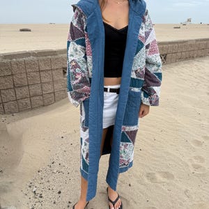 May include: A long, open-front coat featuring a patchwork design of various floral and geometric fabric patterns in shades of blue, white, and maroon. The garment includes a solid blue hood, wide blue lapels, and two large patch pockets.