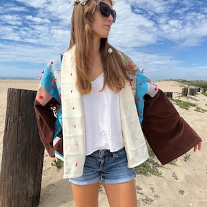 May include: A patchwork kimono-style jacket featuring brown, blue, and grey fabric panels with red pom-pom accents and a cream-colored collar. The jacket is worn over a white camisole and blue denim shorts, paired with a white floral crown and sunglasses.
