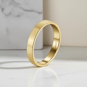 May include: A polished gold-tone band ring with a smooth, flat surface and a rounded profile. The interior of the ring is clearly engraved with the text S925, indicating the metal composition. The ring is captured in a clear, focused view.