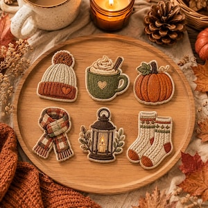Op de afbeelding: Een set van zes geborduurde patches met herfstthema's: een beige en oranje gebreide muts, een groene mok warme chocolademelk met slagroom, een oranje pompoen, een sjaal met ruitjespatroon, een bruine lantaarn en een paar beige gebreide sokken.