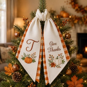 May include: A cream-coloured fabric tree topper featuring two hanging tails with orange and white gingham borders. The left tail displays an embroidered letter T and a pumpkin, while the right tail features the text Give Thanks and a pumpkin.