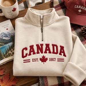 May include: A cream-coloured quarter-zip sweatshirt featuring the word CANADA embroidered in bold red letters across the chest. Below the text, a red maple leaf is centred between the embroidered text EST. 1867 and two horizontal red lines.