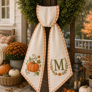 May include: A cream-colored fabric door hanger featuring an orange and white gingham border. One side displays an embroidered orange pumpkin with green vines, while the other side features a large green letter M surrounded by a floral wreath.