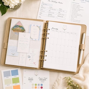 Puede incluir: Un planificador de cuero beige de seis anillas abierto en una página de calendario de junio de 2026, con varios insertos de planificación de bodas, muestras de invitaciones, una tarjeta de paleta de colores, guía de fuentes y un bolígrafo blanco.