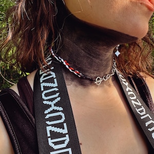 May include: A necklace featuring a braided cord in white, red, and black, connected to a silver-toned metal chain with a toggle clasp. The necklace is worn alongside wide black straps featuring repeating white text that reads KIZUXOZDRO.