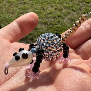 May include: A small opossum figurine crafted from beads, featuring a large spherical body covered in silver faceted crystals, a white pearlescent head with black bead eyes, black bead legs with pink star-shaped feet, and a long gold-toned tail.