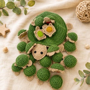 May include: A large crocheted green turtle with a tan head and black eyes, featuring a shell adorned with several smaller crocheted turtles in green, tan, yellow, and lime, all crafted with textured yarn and detailed stitching throughout.