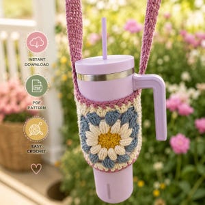 May include: A light purple tumbler with a handle and straw, encased in a crocheted sleeve featuring a multi-colored daisy motif in yellow, cream, and blue. The sleeve includes a matching crocheted strap for carrying. Text includes INSTANT DOWNLOAD, PDF PATTERN, and EASY CROCHET.