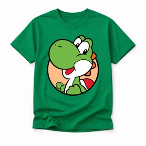 May include: A vibrant green short-sleeved t-shirt featuring a circular graphic print of a green dinosaur character with a white snout, red saddle, and orange spikes on its back, set against a light peach-coloured circular background.