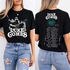 May include: A black t-shirt featuring a graphic of Luke Combs playing an acoustic guitar on the front with the text Luke Combs. The back displays the text Luke Combs, My Kinda Saturday Night Tour, a signature, and a list of tour dates and locations.