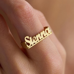 May include: A gold-toned metal ring featuring the name Sienna in a cursive script font. The ring has a flat, horizontal bar design that sits across the finger, with the letters connected to the band in a polished, reflective metallic finish.