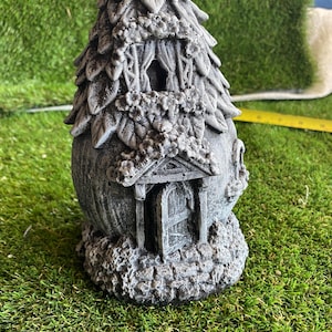 May include: A grey stone-effect decorative house sculpture featuring a textured leaf-patterned roof, a small arched window with curtains, and an open front door. The base is detailed with a cobblestone texture, and a yellow measuring tape is visible.