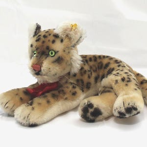 May include: A plush leopard toy featuring tan fur with black spots, bright green eyes, a pink nose, and a red ribbon tied around its neck. The stuffed animal is in a lying position with its tail extended and dark paw pads visible on its feet.